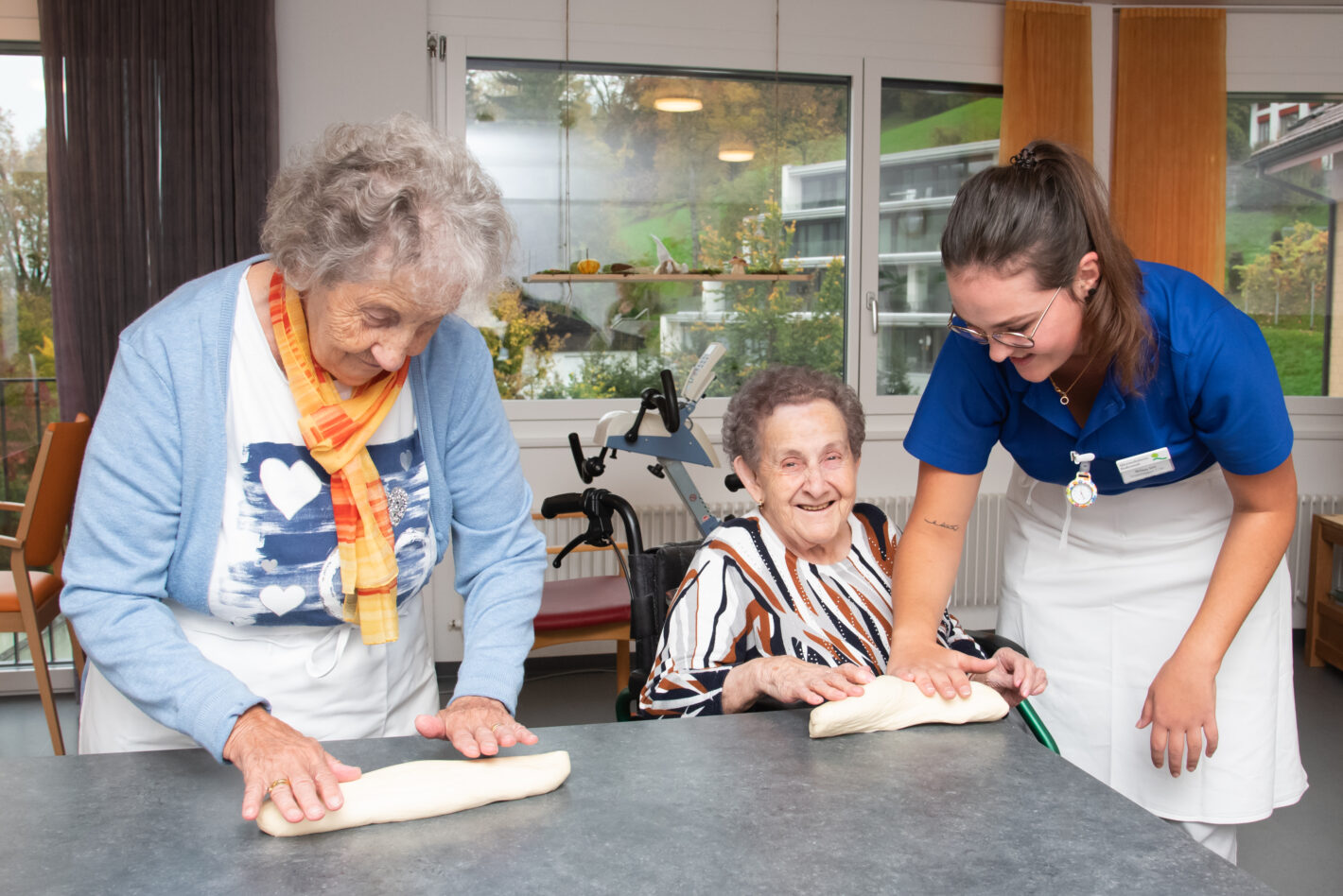 Eine Pflegende hilft zwei Bewohnerinnen den Zopfteig auf der Küchentheke auszurollen.