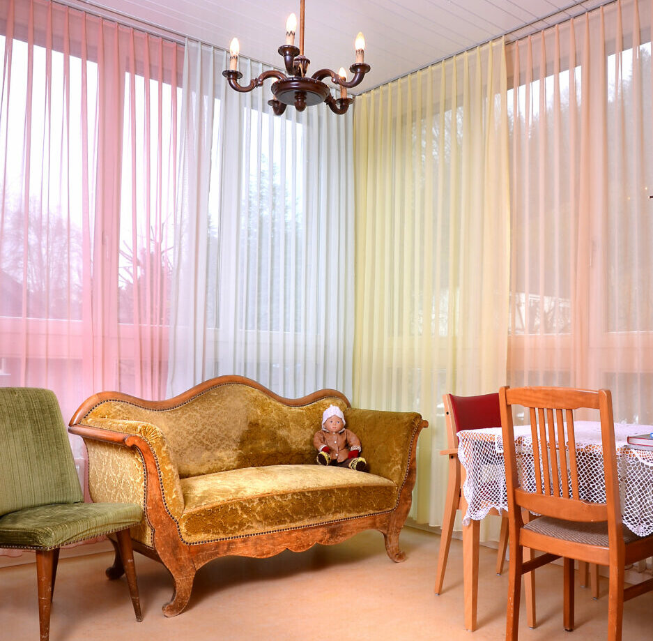 Zimmer für die Bewohner mit einer Demenzerkrankung. Zusehen ist ein Vintage-Sofa mit einer Puppe darauf. Die Vorhänge sind in verschiedenen Pastellfarben.
